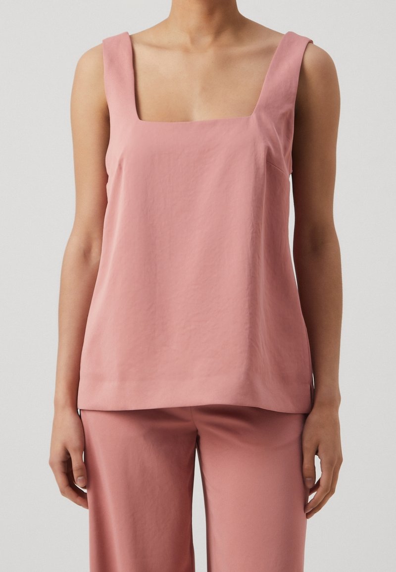 Femme portant un haut rose sans manches à col carré et un pantalon rose ample assorti, debout devant un fond clair uni.