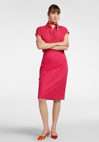 Abito camicia fucsia con maniche corte, colletto abbottonato, vita aderente, gonna di lunghezza ginocchio e tacchi a punta arancioni. Accento di sciarpa.