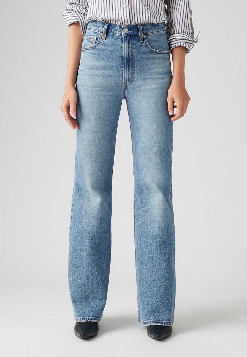 Femme portant un jean droit taille haute bleu clair, associé à des chaussures noires pointues et une chemise blanche rayée aux manches retroussées.