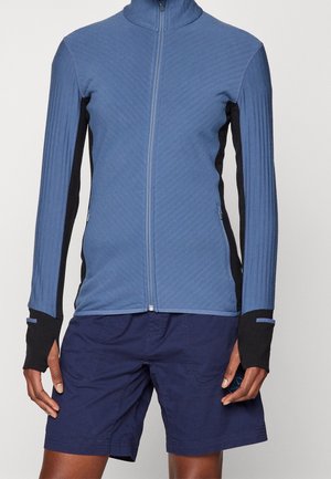 Veste bleue zippée avec panneaux latéraux noirs et poignets avec trou pour le pouce, portée avec un short bleu marine.