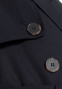 Mörkblått tygerockdetalj med en stor brun knapp med fyra hål och en vikbar krage, som visar upp en slät textur och skräddarsydd design.