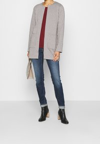 Manteau long à carreaux crème et noir, devant ouvert, avec deux poches, associé à un haut bordeaux, un jean bleu et des bottines noires à talons.