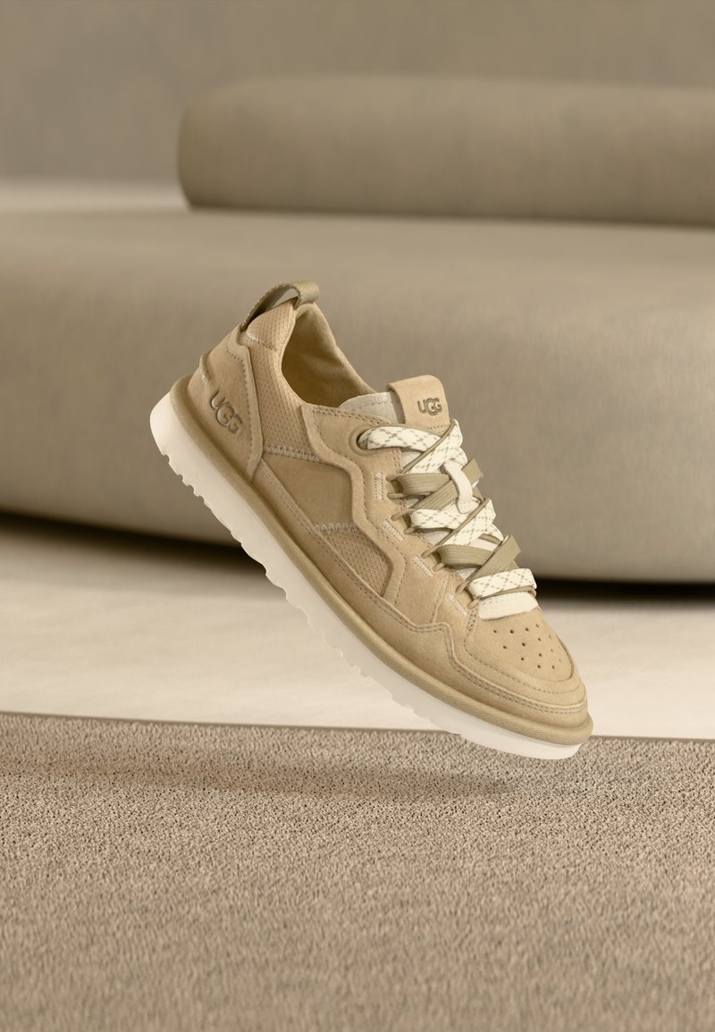 Baskets en daim beige avec semelle blanche texturée, dotées d'un bout arrondi, d'un laçage avant et de la marque UGG. Les accents comprennent des lacets gris clair.