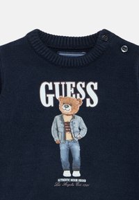 Μπλε ναυτικό πλεκτό πουλόβερ με γραφικό αρκούδας που φοράει τζιν μπουφάν. Κείμενο «GUESS» πάνω και κείμενο «AUTHENTIC DENIM BRAND» κάτω.
