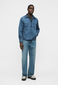 Muž oblečený v modrej vetrovke na zips, čiernom tričku pod ňou, svetlomodrých rifle so širšími nohavicami a béžových topánkach s čiernou podrážkou, stojaci s jednou rukou v vrecku.