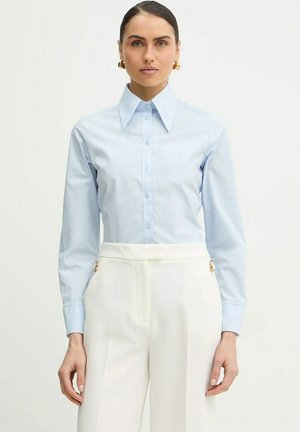 Donna che indossa una camicia azzurro chiaro con colletto appuntito e pantaloni bianchi a vita alta con dettagli di fibbie dorate.