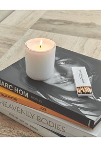 Candela bianca accesa e scatola di fiammiferi sopra un mucchio di quattro libri d'arte e fotografia su una superficie piastrellata.