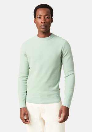 Mies, joka pukeutunut mintunvihreään ribbineulottuun pitkähihaiseen villapaitaan ja valkoisiin housuihin, seisoo yksinkertaisen vaalean taustan edessä.