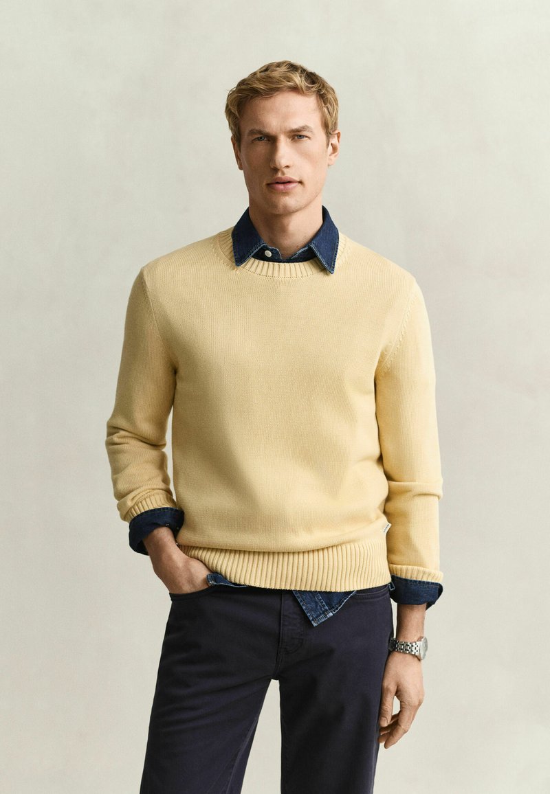 Homme portant un pull jaune clair sur une chemise en jean bleu foncé avec un pantalon foncé, debout avec une main dans la poche devant un fond uni.