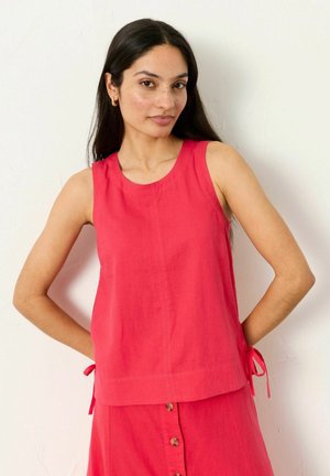 Naine, kellel on pikad tume juuksed ning kes kannab kuldseid rõngakujulisi kõrvarõngaid, varrukateta punast topi külgtanssidega ja sobivaid punaseid nööpidega seelikuid, seisab käed selja taga.