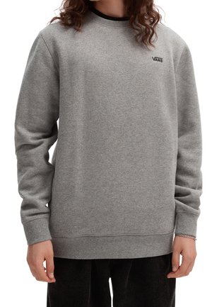 Personne portant un sweat à capuche gris de Vans avec des manches longues, des poignets côtelés et un pantalon noir, se tenant devant un fond blanc.