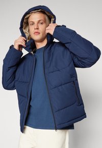 Giacca puffer blu navy con cappuccio, texture trapuntata, zip nera e rivestimento beige. Il modello sta regolando il cappuccio mentre indossa una camicia blu.