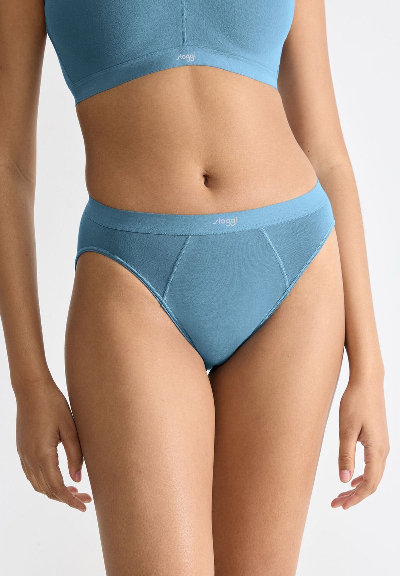 Donna che indossa un completo coordinato di biancheria intima e reggiseno sportivo in cotone azzurro chiaro, in piedi contro uno sfondo chiaro e semplice.