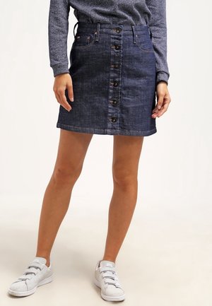 Vrouw draagt een donkerblauwe spijkerrok met knopen aan de voorkant, grijs shirt met lange mouwen en witte sneakers, staat met benen iets uit elkaar.