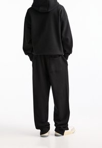 Schwarzer Kapuzenpullover mit einem cropped Design, kombiniert mit lockeren Hosen mit einer Gesäßtasche und beigen Sneakern mit einer hellen Sohle.