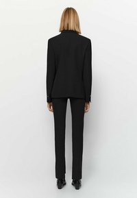 Veste de costume noire ajustée avec une coupe slim, associée à un pantalon tailleur assorti. Elle présente une fente unique et des éléments de design minimalistes.