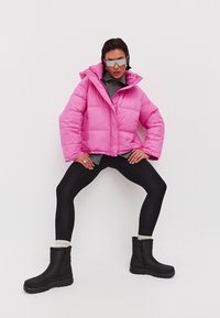 Chaqueta de plumas rosa con un cuello alto, combinada con una camiseta gris y leggings negros. Botas de invierno negras con una superficie texturizada y cremallera.