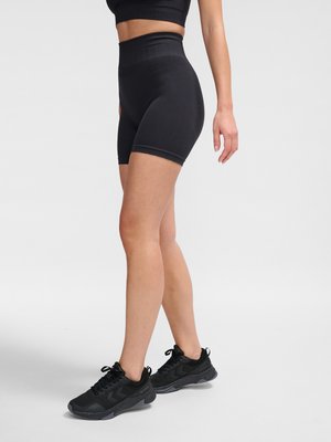 Kobieta nosząca czarne rowerowe szorty z wysokim stanem, czarny krótki top i czarne buty sportowe, stojąca ze skrzyżowanymi nogami na białym tle.