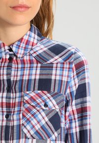 Rutig skjorta i marinblått, röd och blått. Har en klassisk krage, knappstängning fram och en vänster bröstficka med diagonalt lock. Mjuk textur.