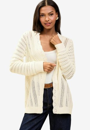Strickjacke in Creme mit Lochmustern, langen Ärmeln und gerippten Bündchen. Das Modell trägt ein weißes Tanktop und dunkle Jeans.