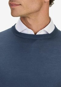 Blå stickad tröja med rund halsringning, med en mjuk textur. Bärs över en vit skjorta med krage och knappstängning.