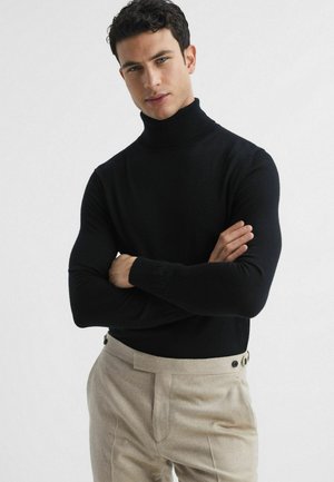 Pulover negru cu guler înalt, realizat dintr-un material moale, cu un design ajustat și mâneci lungi. Asortat cu pantaloni bej cu accente de nasturi.