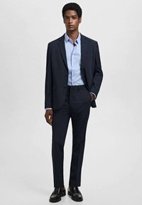 Donker marineblauw op maat gemaakt pak met een blazer met één knoop, gecombineerd met een lichtblauw overhemd en zwarte nette schoenen. Slim-fit broek en klassiek ontwerp.