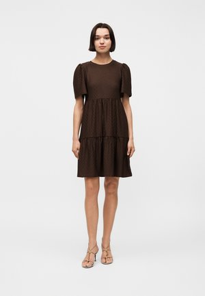 Žena s krátkymi tmavými vlasmi, ktorá má na sebe tmavohnedé šaty po kolená s nadýchanými krátkymi rukávmi a béžové vysoké sandále so remienkami.