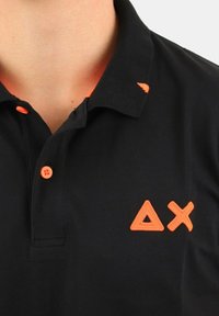 Polo nero con bottoni arancioni e dettagli di logo geometrico ricamato; presenta un colletto e un tessuto testurizzato.