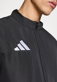 Μαύρο μπουφάν με ψηλό γιακά, φερμουάρ στο μπροστινό μέρος και λευκό κεντητό logo με τρεις ρίγες στο αριστερό στήθος. Ομαλή υφή.