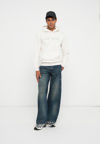 Sudadera blanca de algodón con el logo "GUESS", combinada con pantalones vaqueros de pierna ancha en azul que presentan detalles de costura y zapatillas grises. Se lleva una gorra negra.