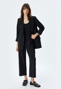 Zwarte blazer met een ontspannen pasvorm, gecombineerd met een zwarte aansluitende tanktop en wijde broek. Het materiaal lijkt glad; de schoenen hebben een gestructureerde afwerking.