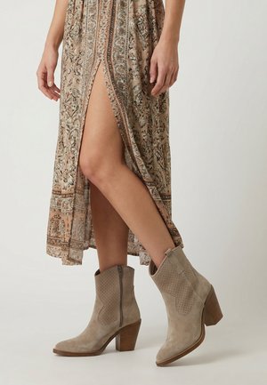 Kvinde iført beige ankelstøvler i ruskind med perforerede detaljer og en mellemhøj blokhæl, kombineret med et mønstret midi-nederdel med frontslids.