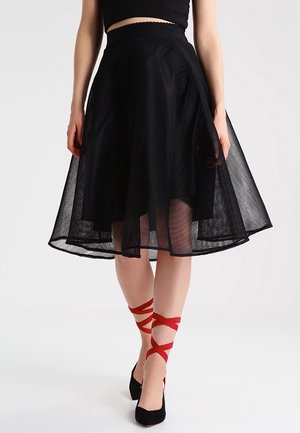 Kobieta ubrana w czarną, wysoką siateczkową spódnicę, czarną bluzkę, czarne szpilki oraz czerwone krzyżujące się wstążki na kostkach, stojąca na tle jednolitym.