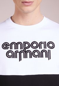 White and black cotton shirt featuring a large embroidered "emporio armani" logo in black across the chest with a rounded neckline.