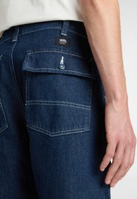 Los pantalones de mezclilla azul oscuro tienen bolsillos traseros rectangulares, costuras blancas y una pequeña etiqueta de logo con un gráfico. Detalle de botón en los bolsillos.