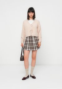 Cardigan court rose clair avec un fermeture à nouer, associé à une mini-jupe à carreaux marron. Des chaussettes beiges transparentes et des ballerines bordeaux complètent le look.