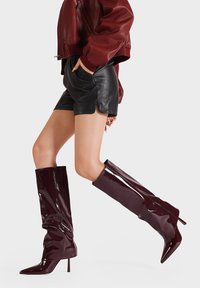 Stivali alti in pelle verniciata bordeaux con design a sbuffo, punta a mandorla e fitting fino al ginocchio. Texture liscia e finitura lucida.