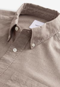 Beige overhemd met knoopsluiting, gemaakt van textuurstof, met een standaard kraag, twee knopen bij de kraag en een zak aan de voorkant.