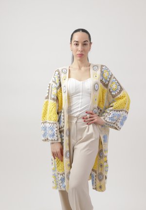 Vrouw staand met hand op haar heup, gekleed in een kleurrijke patchwork gebreide cardigan, een witte korset top en beige broek met hoge taille tegen een effen achtergrond.