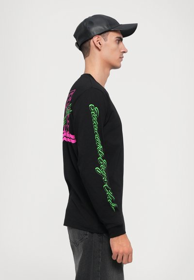 Camisa preta de manga longa com gráficos vibrantes em rosa e verde na frente e nas mangas, combinada com um boné de couro preto.