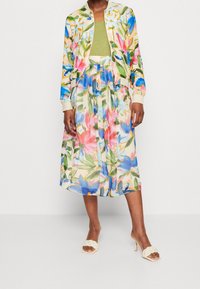 Blomstret bomberjakke i let stof, med pastel farver og livlige mønstre; parret med en grøn top og en gennemsigtig, svævende nederdel.