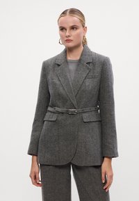 Sivi herringbone sako s krojem koji prati tijelo, jednostruko zakopčavanje, reveri sa zarezom i strukiranim pojasom. Ima dva prednja džepa.