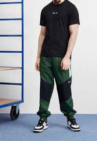 Svart t-shirt med "A I R"-logotyp, ihop med gröna och svarta panelbyxor. Modellen bär svarta och vita sneakers. Blå bakgrund.