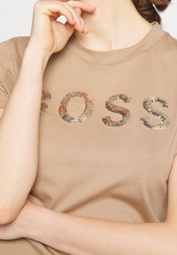 Beżowa bawełniana koszulka z krótkim rękawem, z ozdobnymi literami w kolorze złotym i srebrnym, które tworzą napis "BOSS" na piersi.