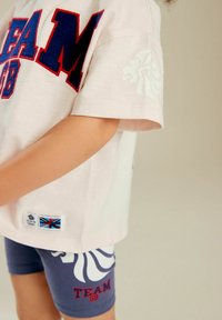 Helles pinkes T-Shirt mit roter und blauer "TEAM GB" Schrift. Marineblaue Shorts mit einem weißen Löwe-Graphic und rotem "TEAM GB" Text.