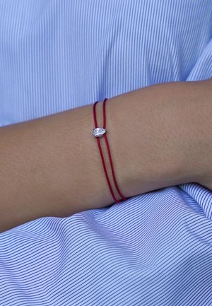 Bras portant un bracelet double brin rouge avec une pierre claire en forme de goutte sur un fond en tissu rayé bleu et blanc.