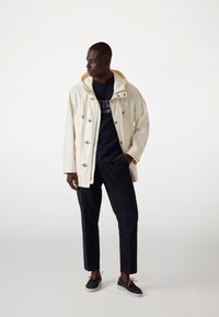 Witte, capuchonjack met sluitingen, gedragen over een marineblauw t-shirt en donkere broeken. Leren schoenen met bruine veters en lichte zolen.