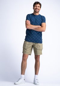 Tricou bleumarin cu model de puncte verzi, pantaloni scurți cargo bej, adidași albi și șosete albe. Ținuta are un croi lejer, casual.