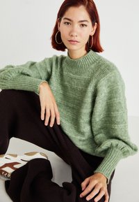 Light green knitted sweater with a ribbed turtleneck, featuring loose sleeves. Dark brown textured pants and white sneakers with brown accents.
